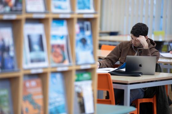 una persona che studia con appunti e un computer portatile, e uno scaffale di riviste in primo piano